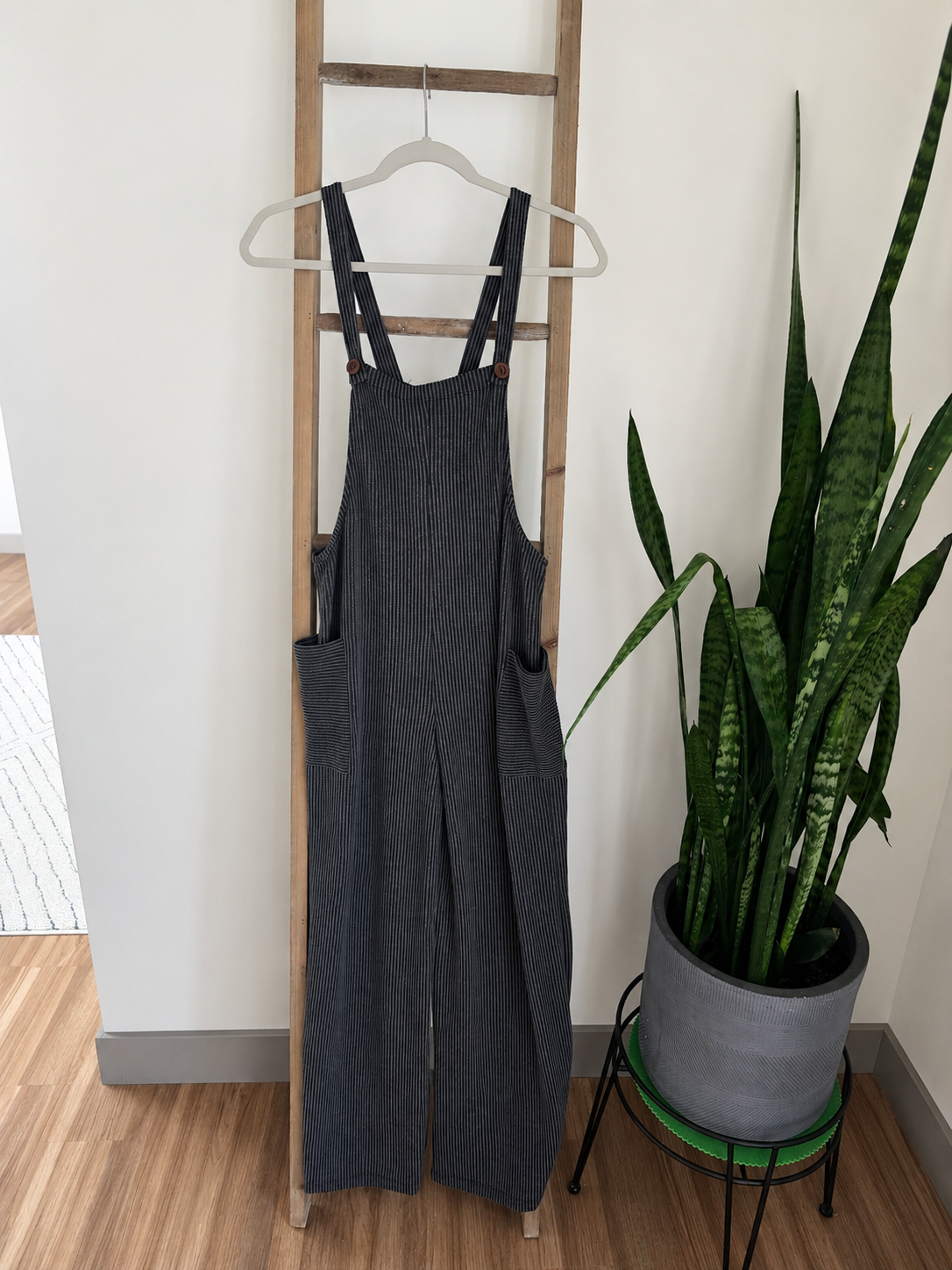 Dark gray corduroy bib overalls hanging on a wooden hanger on a wooden ladder, next to a potted snake plant in a home setting.