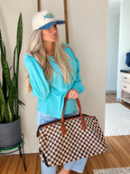 A woman stands indoors holding a large checkered duffel bag with brown leather handles. She is wearing a turquoise sweatshirt, light blue jeans, a white cap with a blue patterned brim, and a gold necklace.