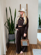 A woman stands barefoot in a room wearing a dark brown cropped sweatshirt and matching wide-leg pants, holding a brown handbag with a black-and-white checkered front panel. She is wearing a white baseball cap with leopard-print trim.