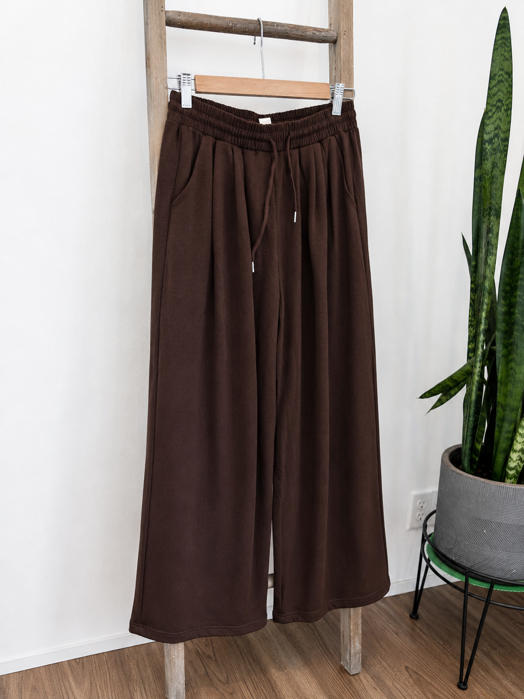 A pair of dark brown wide-leg sweatpants hanging from a wooden hanger on a wooden ladder, with a potted snake plant in the background.