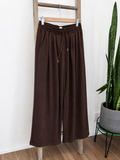 A pair of dark brown wide-leg sweatpants hanging from a wooden hanger on a wooden ladder, with a potted snake plant in the background.