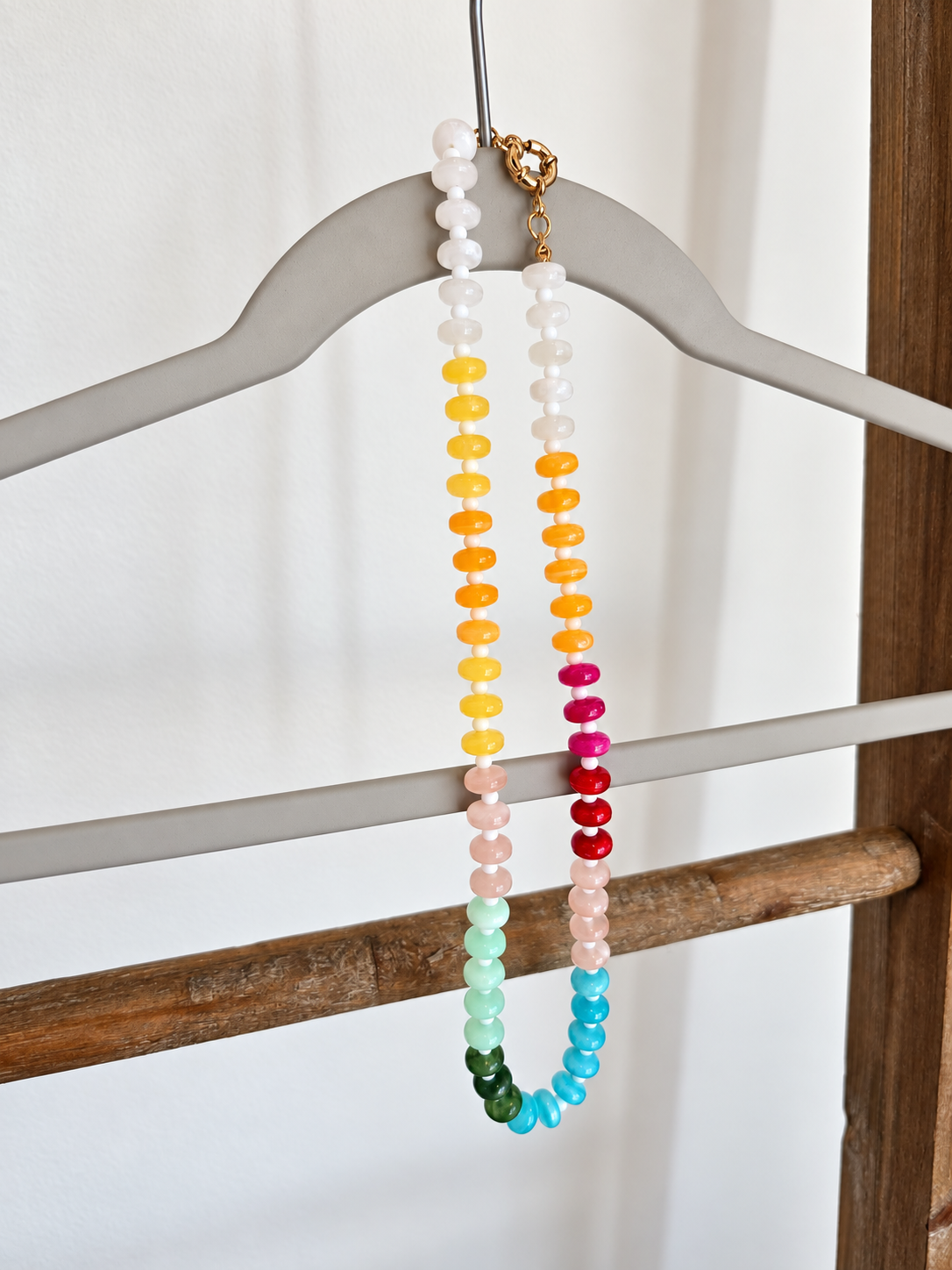 A beaded necklace with multiple strands in rainbow colors hangs from a gray wooden hanger. The necklace features flat, disc-like beads in shades of yellow, orange, pink, red, blue, green, and white, with a small gold clasp at the top.