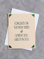 A beige congratulations card with dark green text reading 'CONGRATS ON THE NEW BABY. I KNEW YOU HAD IT IN YOU.