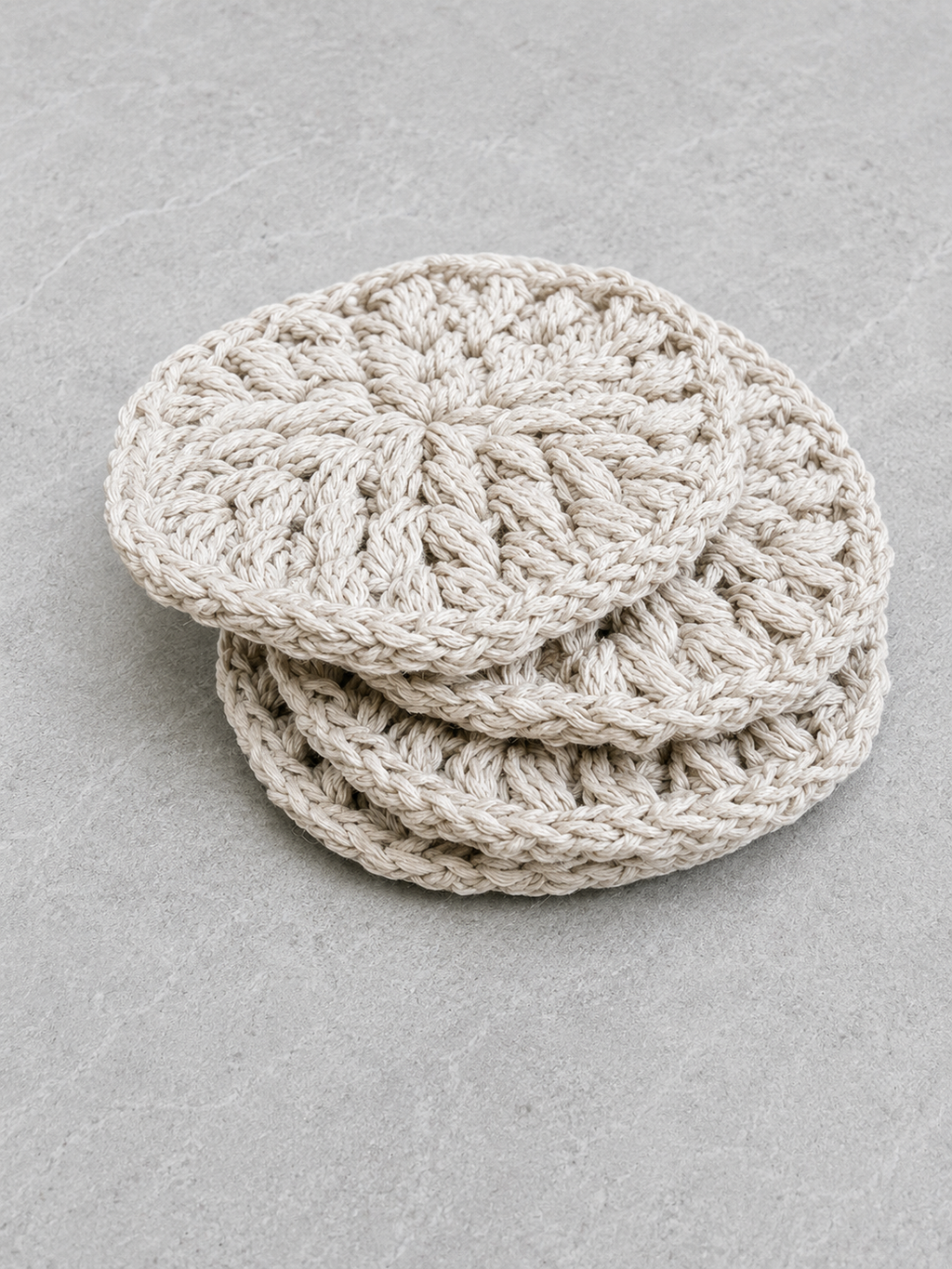 Four round crocheted coasters in a light oatmeal beige color, stacked on a textured gray surface.