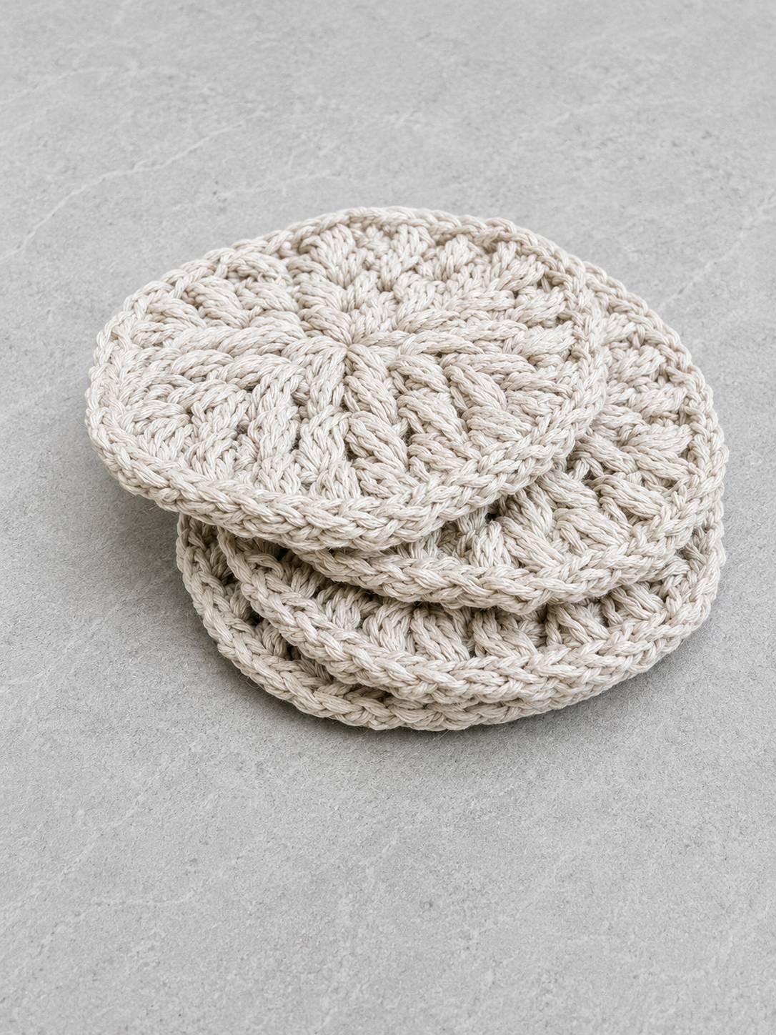 Four round crocheted coasters in a light oatmeal beige color, stacked on a textured gray surface.