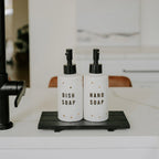 Two white textured pump bottles labeled 'DISH SOAP' and 'HAND SOAP' with black pumps, sitting on a black tray on a white countertop next to a black faucet.