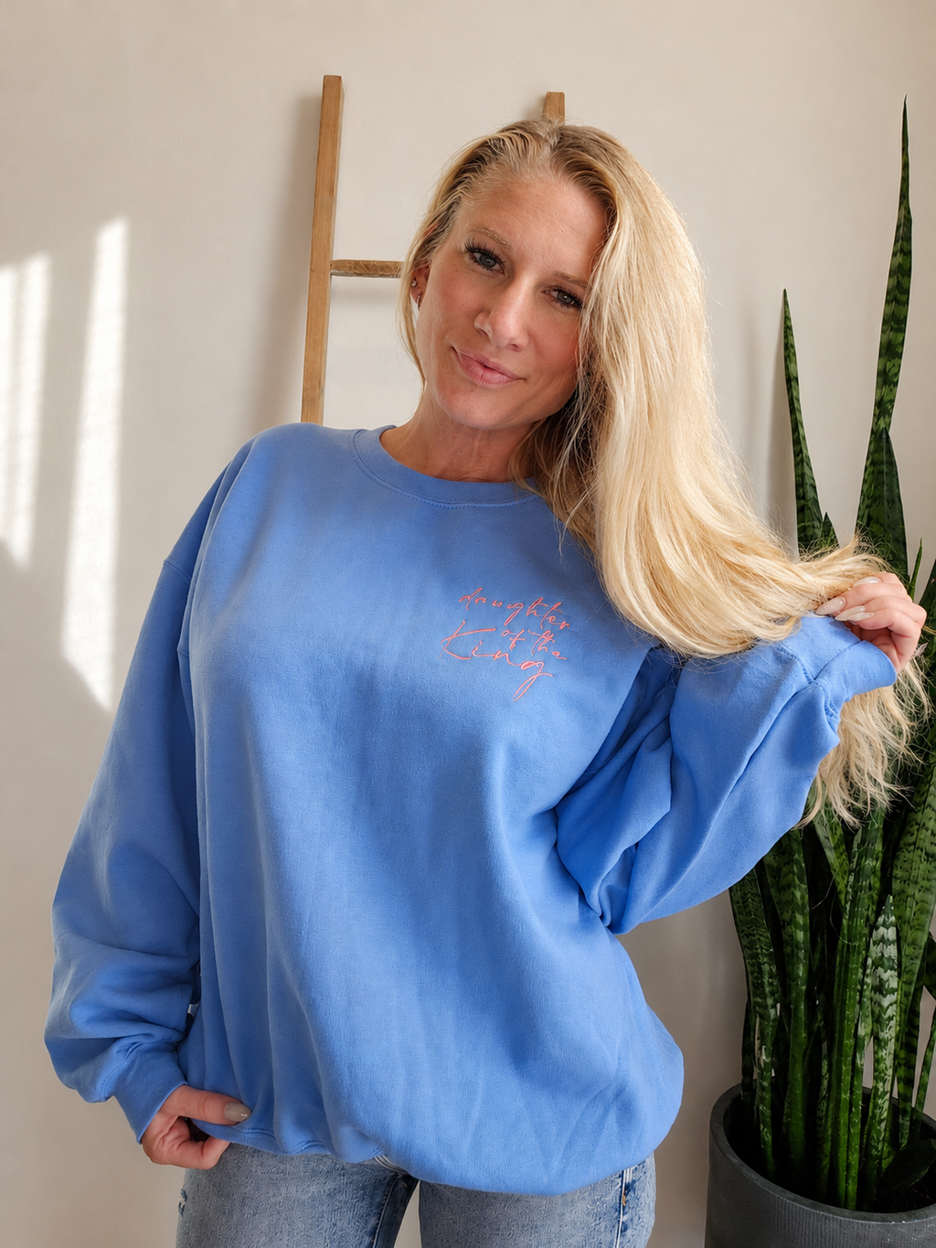 A blonde woman wearing a light blue crewneck sweatshirt with pink script embroidery on the chest, posing in a home setting next to a potted snake plant.