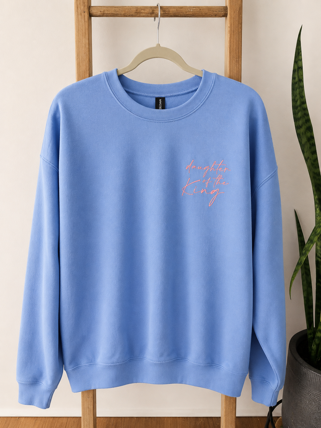 A light blue crewneck sweatshirt hanging on a wooden hanger against a light beige wall, with a small pink script graphic on the left chest. A wooden ladder stand supports the sweatshirt, and a green snake plant is visible on the right side.
