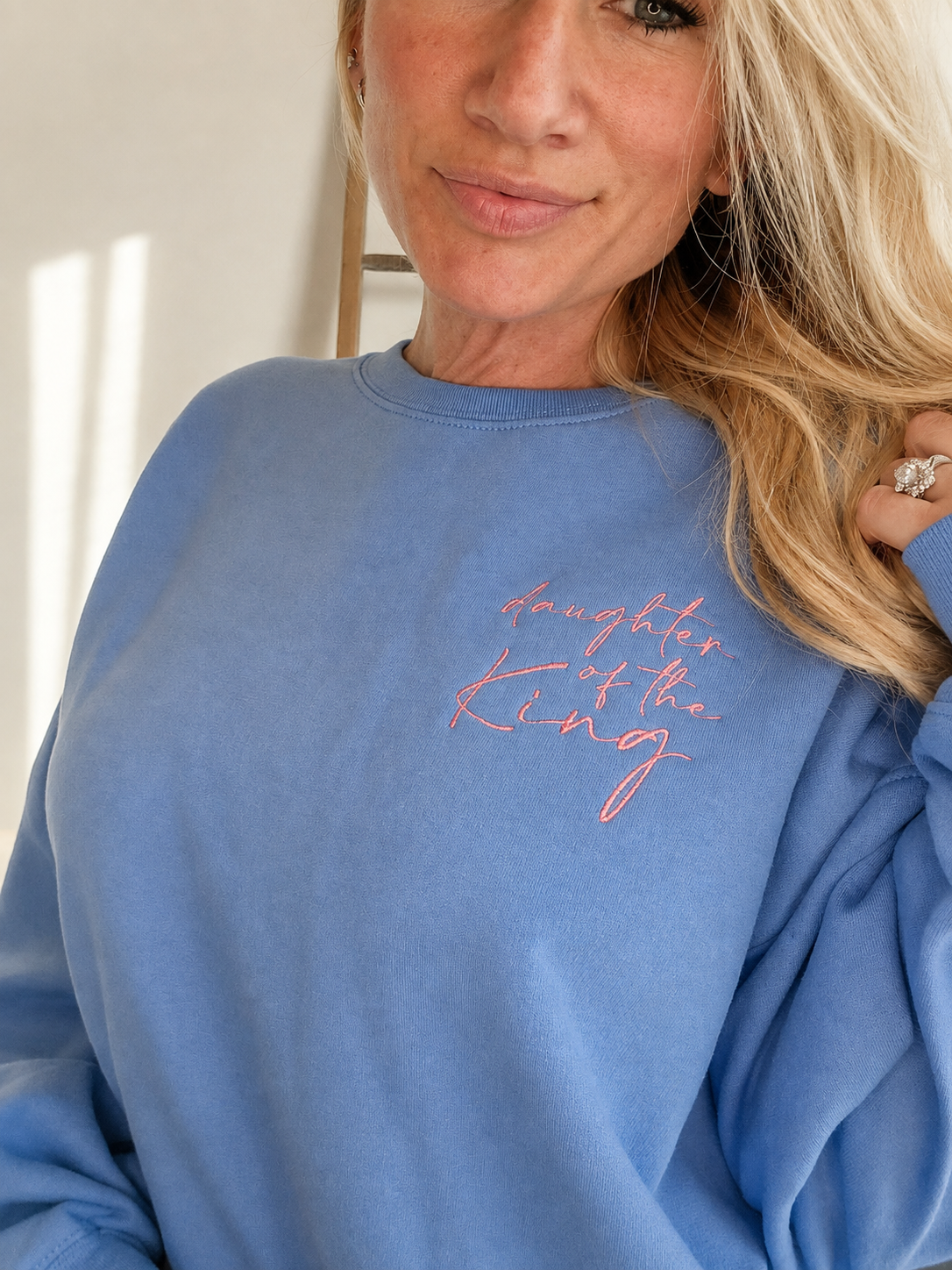 A close-up of a woman wearing a dusty blue crewneck sweatshirt with 'daughter of the king' embroidered in pink script on the chest.