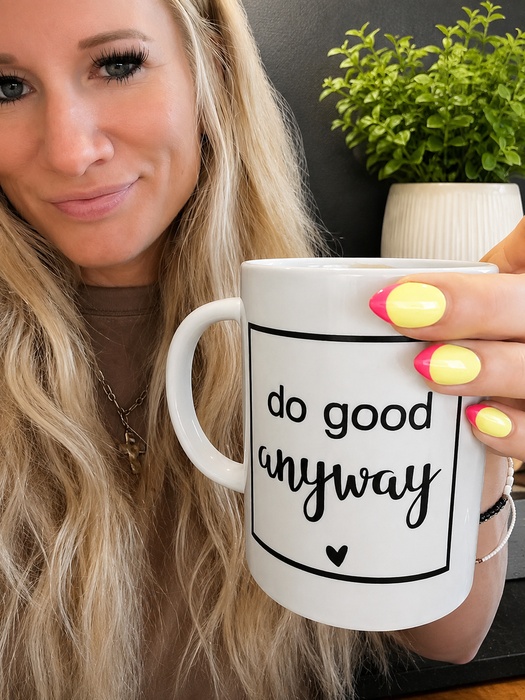 A woman with blonde hair holds a white ceramic mug with the phrase 'do good anyway' printed in black script inside a thin black rectangular border. The background features a dark gray wall and a small potted green plant.