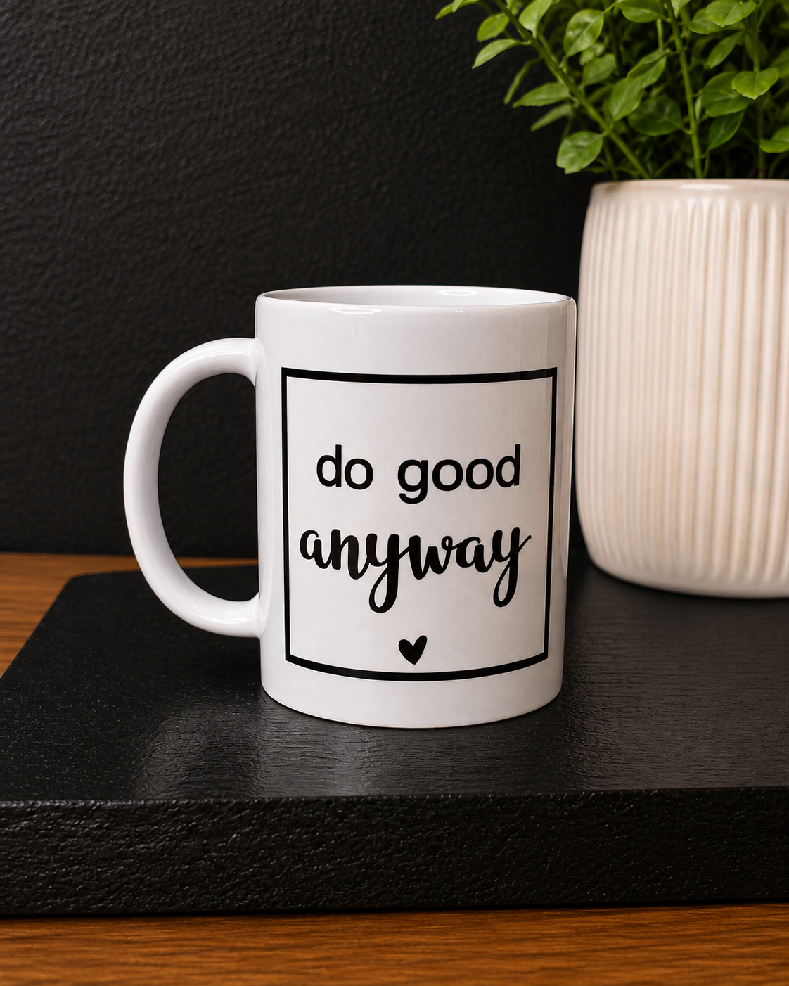 A white ceramic coffee mug with black text reading 'do good anyway' and a small heart, displayed on a black tray with a potted plant in the background.