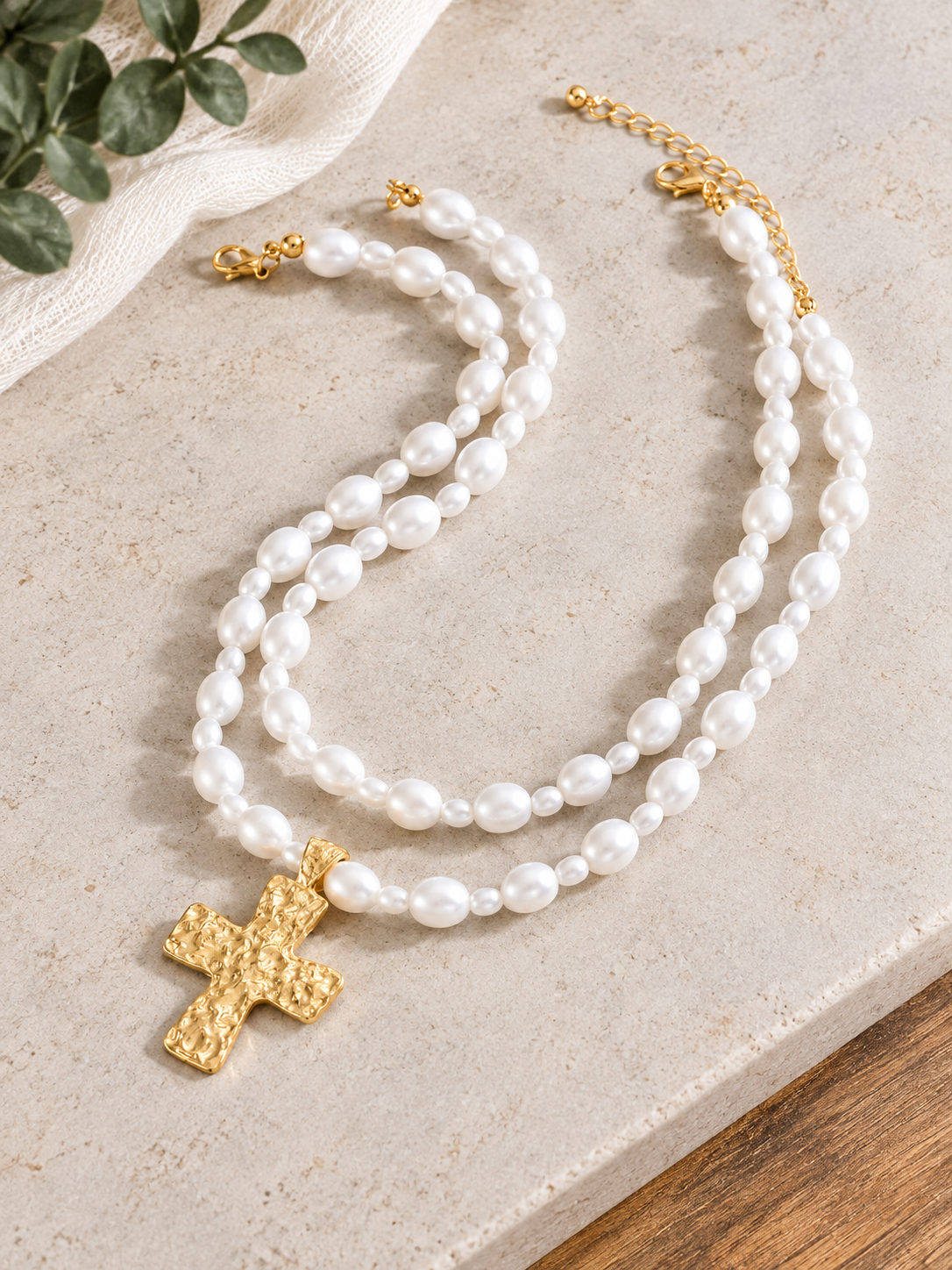 A double-strand pearl necklace with a textured gold cross pendant, displayed on a light stone surface with a small green plant leaf in the corner.