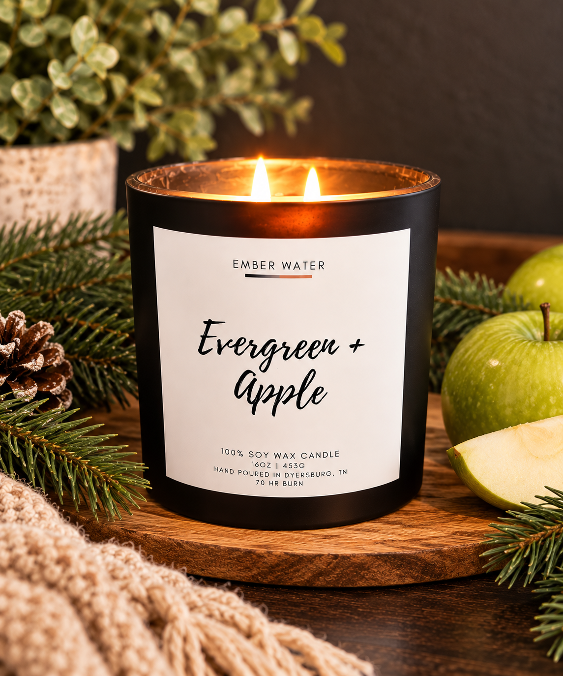 A lit black soy wax candle jar with a white label reading 'EMBER WATER Evergreen + Apple' sits on a wooden board surrounded by green apples, pine branches, a pinecone, and a knitted textile in a styled indoor setting.
