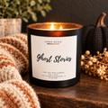 A lit black soy wax candle jar labeled 'EMBER WATER Ghost Stories' sits on a wooden surface with a cozy knit blanket, dried flowers, and a dark pumpkin in the background.