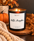 A lit black candle jar labeled 'Hello, Pumpkin' by Ember Water, styled on a wooden board with a small pumpkin, knit blanket, dried flowers, and autumn decor.