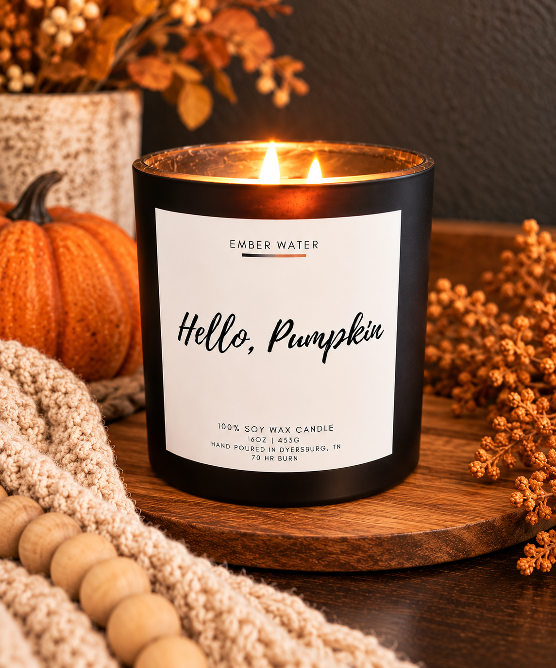 A lit black candle jar labeled 'Hello, Pumpkin' by Ember Water, styled on a wooden board with a small pumpkin, knit blanket, dried flowers, and autumn decor.