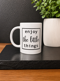 A white ceramic mug with the phrase 'enjoy the little things' printed in black text, styled on a black slate tray next to a potted green plant against a dark textured wall.