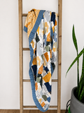 A colorful patchwork quilt with geometric patterns in orange, navy, gray, and white, draped over a wooden ladder next to a potted snake plant.