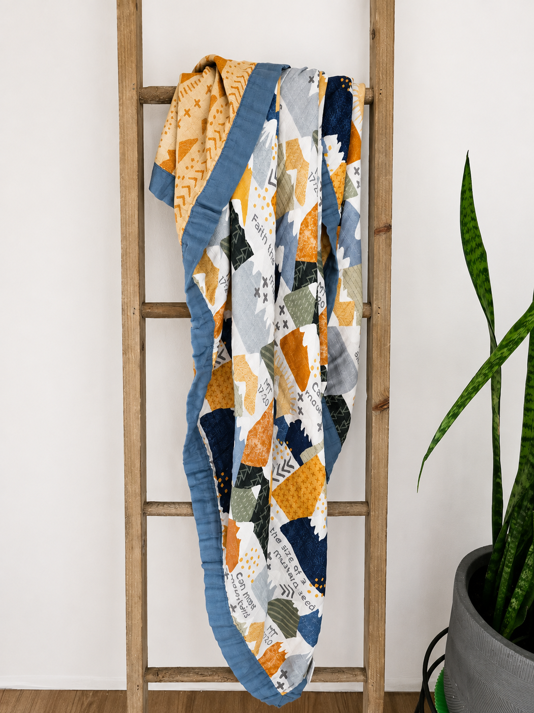 A colorful patchwork quilt with geometric patterns in orange, navy, gray, and white, draped over a wooden ladder next to a potted snake plant.