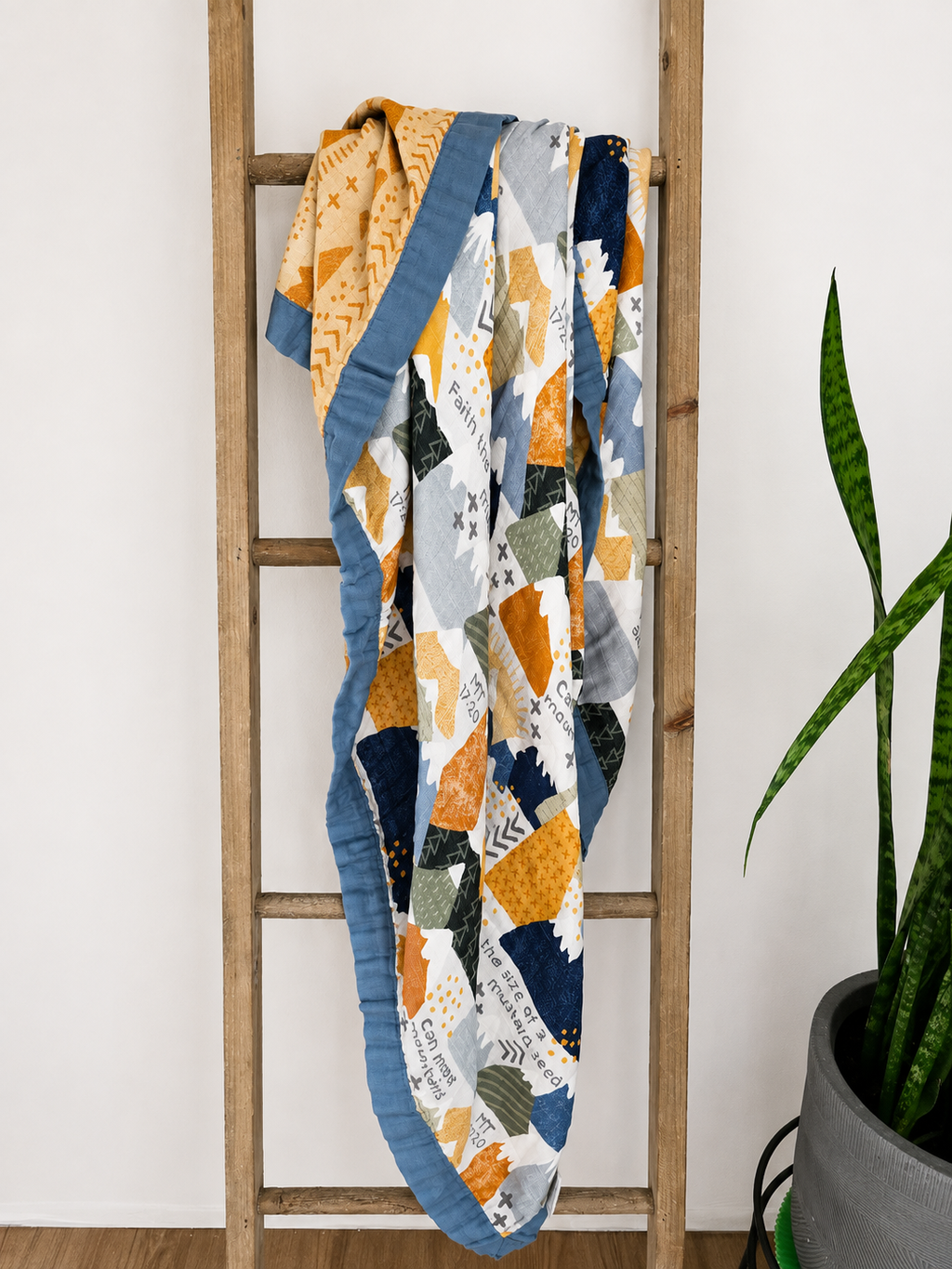 A colorful patchwork quilt with geometric patterns in orange, navy, gray, and white, draped over a wooden ladder next to a potted snake plant.