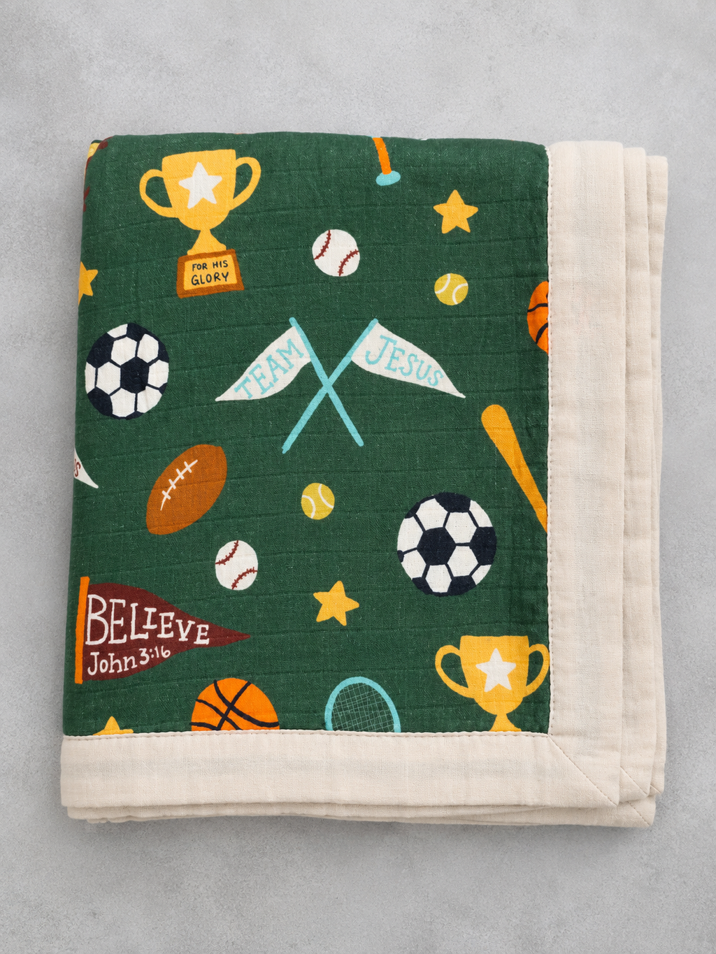 A folded quilted blanket with a dark green sports-themed print featuring soccer balls, basketballs, a football, trophies, stars, and crossed flags, finished with a thick cream-colored border, shown on a light gray background.