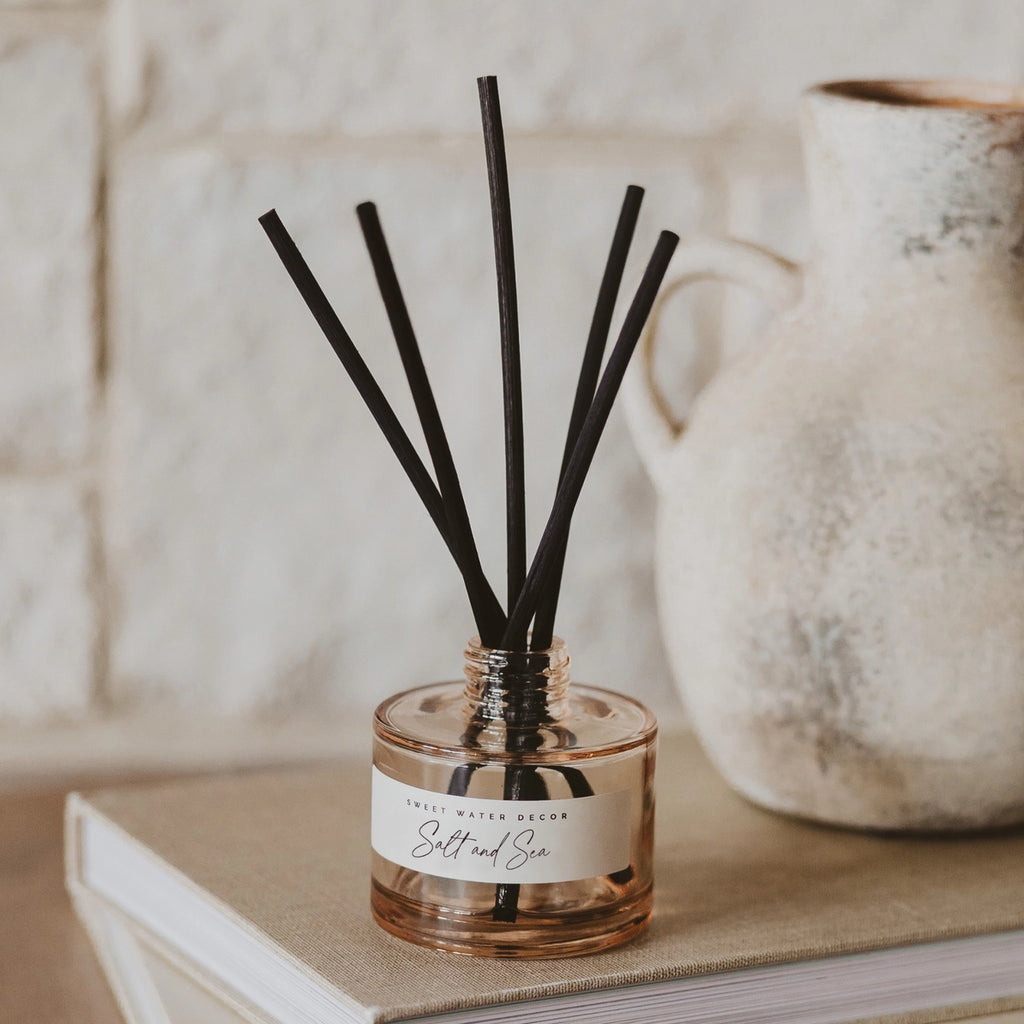 A glass reed diffuser bottle filled with amber-toned liquid, with several black reeds extending upward, placed on a beige book in front of a rustic ceramic pitcher against a light stone wall.
