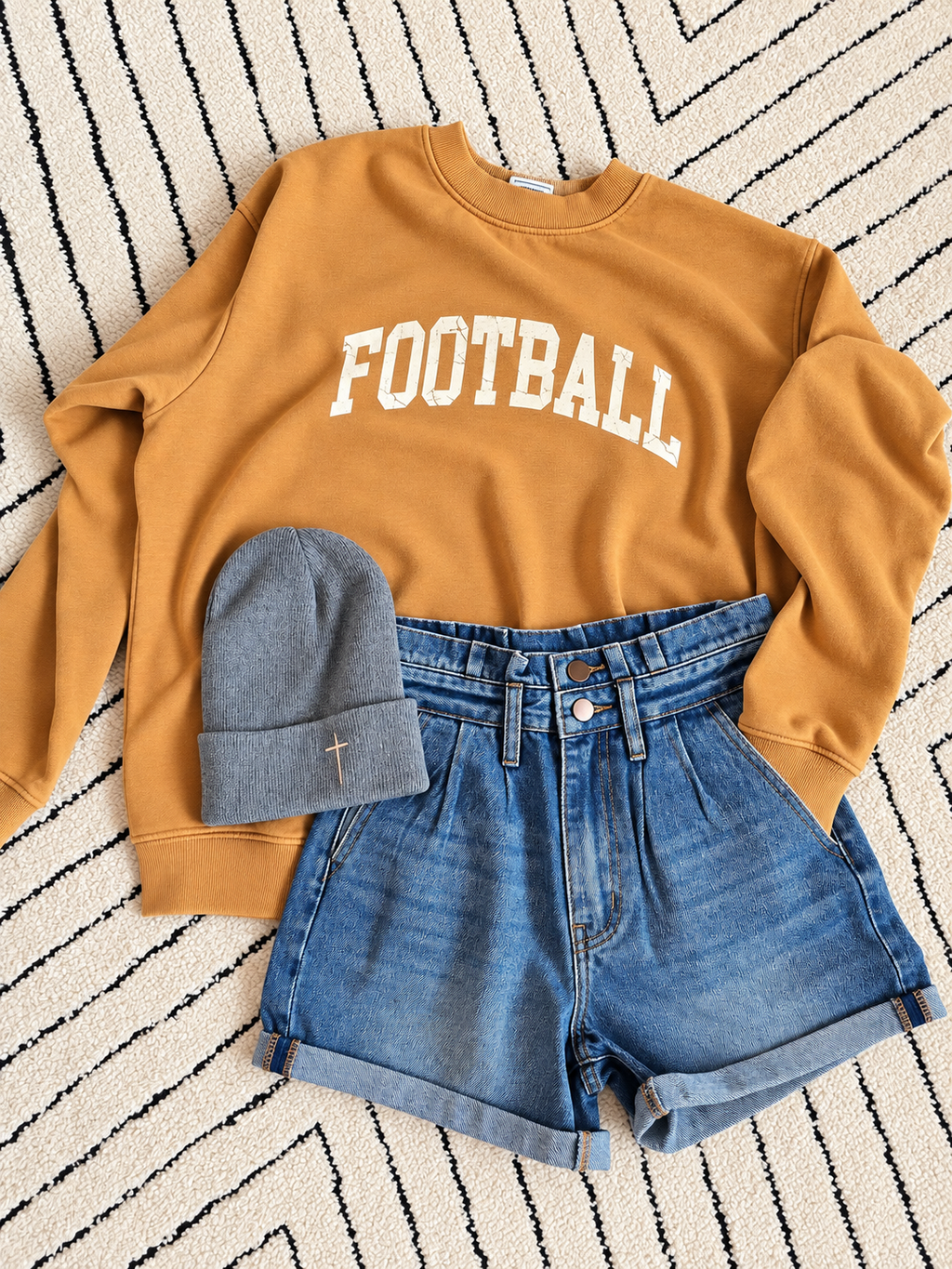 A styled flat-lay outfit featuring a mustard yellow sweatshirt with 'FOOTBALL' printed in white arched letters, paired with blue denim shorts and a gray knit beanie on a black and white patterned rug.