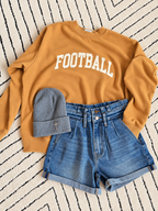 A styled flat-lay outfit featuring a mustard yellow sweatshirt with 'FOOTBALL' printed in white arched letters, paired with blue denim shorts and a gray knit beanie on a black and white patterned rug.