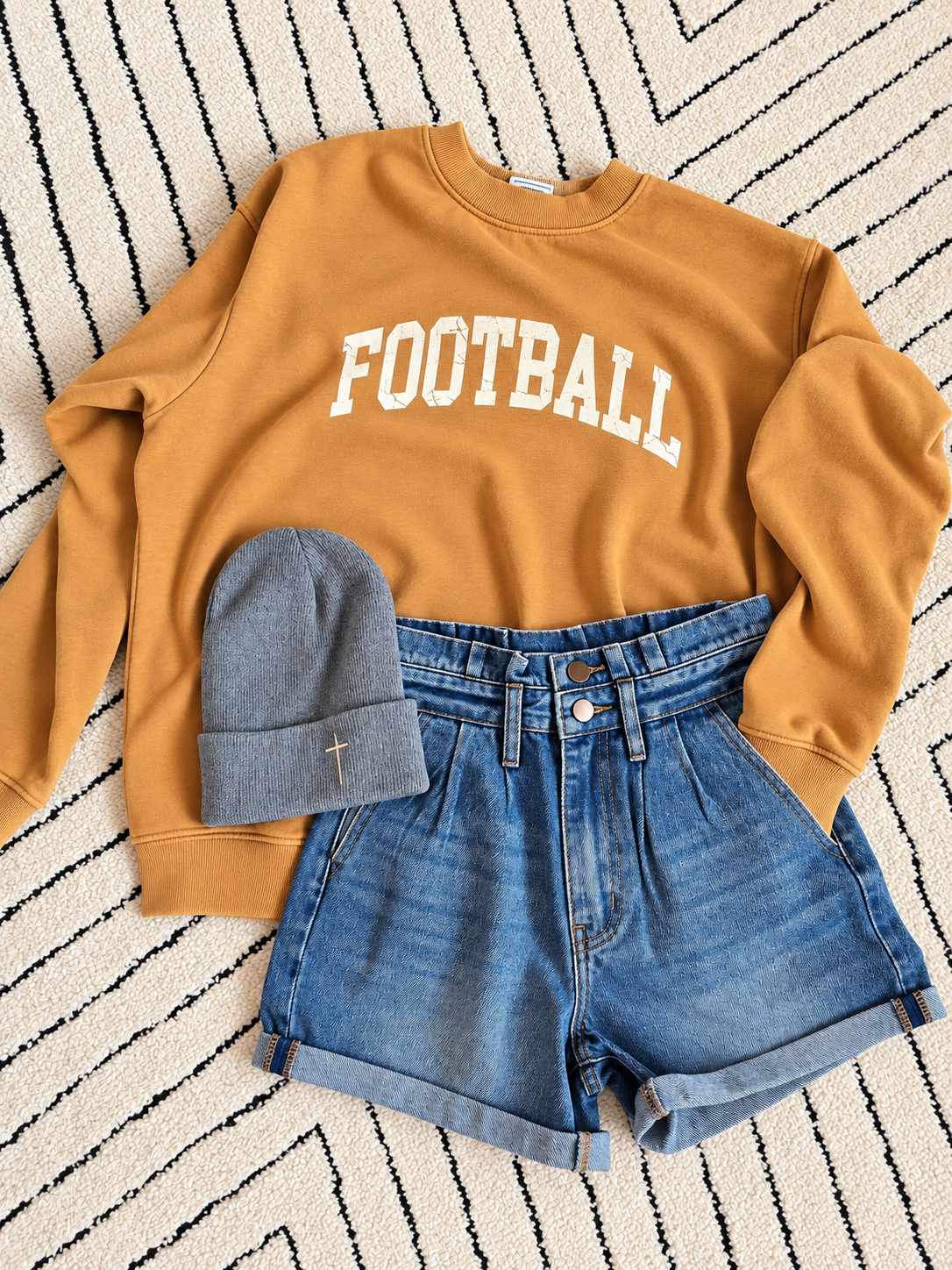 A styled flat-lay outfit featuring a mustard yellow sweatshirt with 'FOOTBALL' printed in white arched letters, paired with blue denim shorts and a gray knit beanie on a black and white patterned rug.