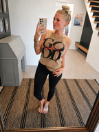 A woman takes a mirror selfie in a home setting, wearing a beige sleeveless tank top with a black graphic print and red bow detail, paired with black leggings.