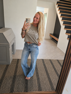 A woman takes a mirror selfie in a home entryway, wearing a light brown cropped t-shirt with a white geometric graphic and light-wash blue flare jeans.
