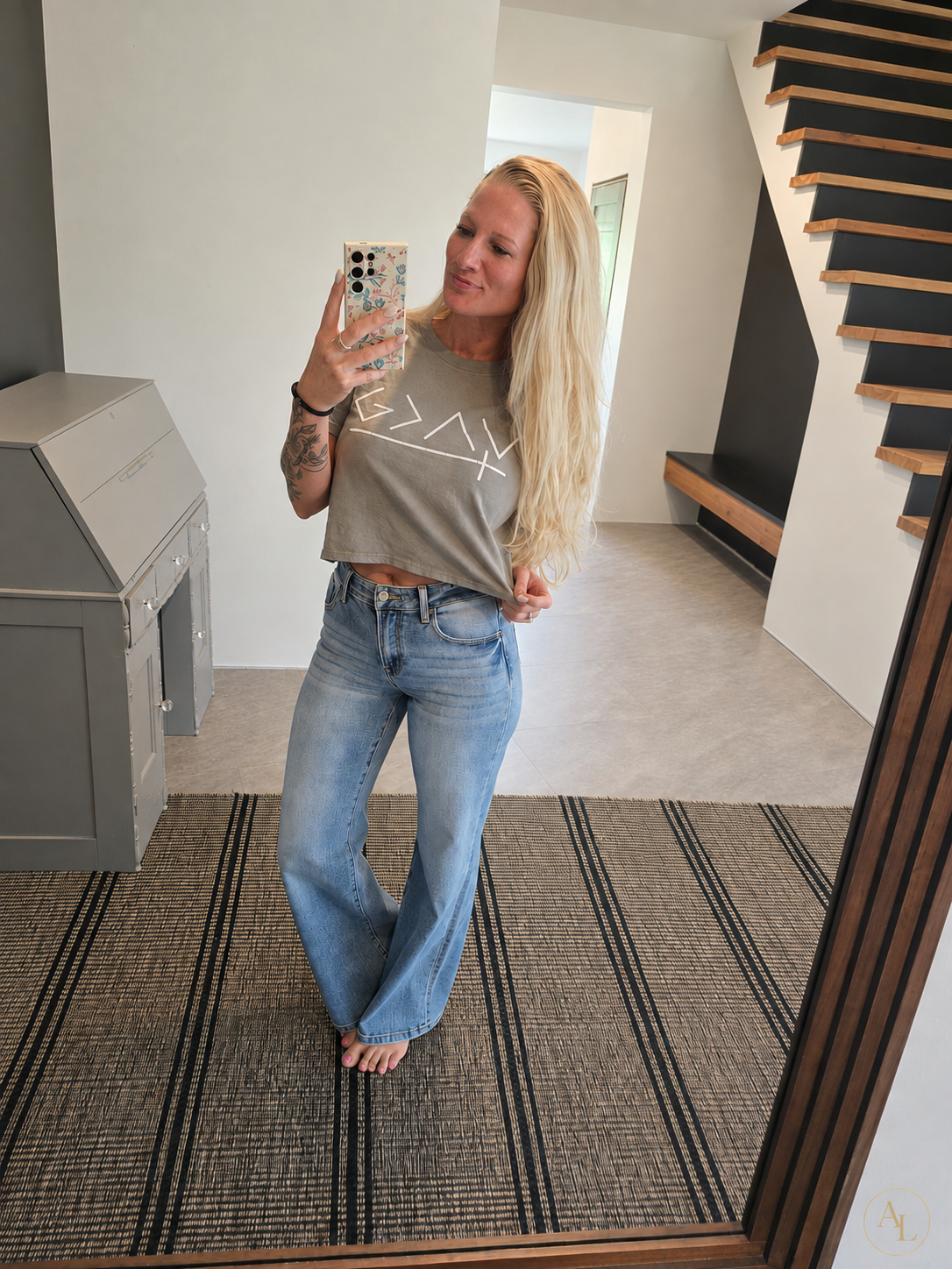 A woman takes a mirror selfie in a home entryway, wearing a light brown cropped t-shirt with a white geometric graphic and light-wash blue flare jeans.