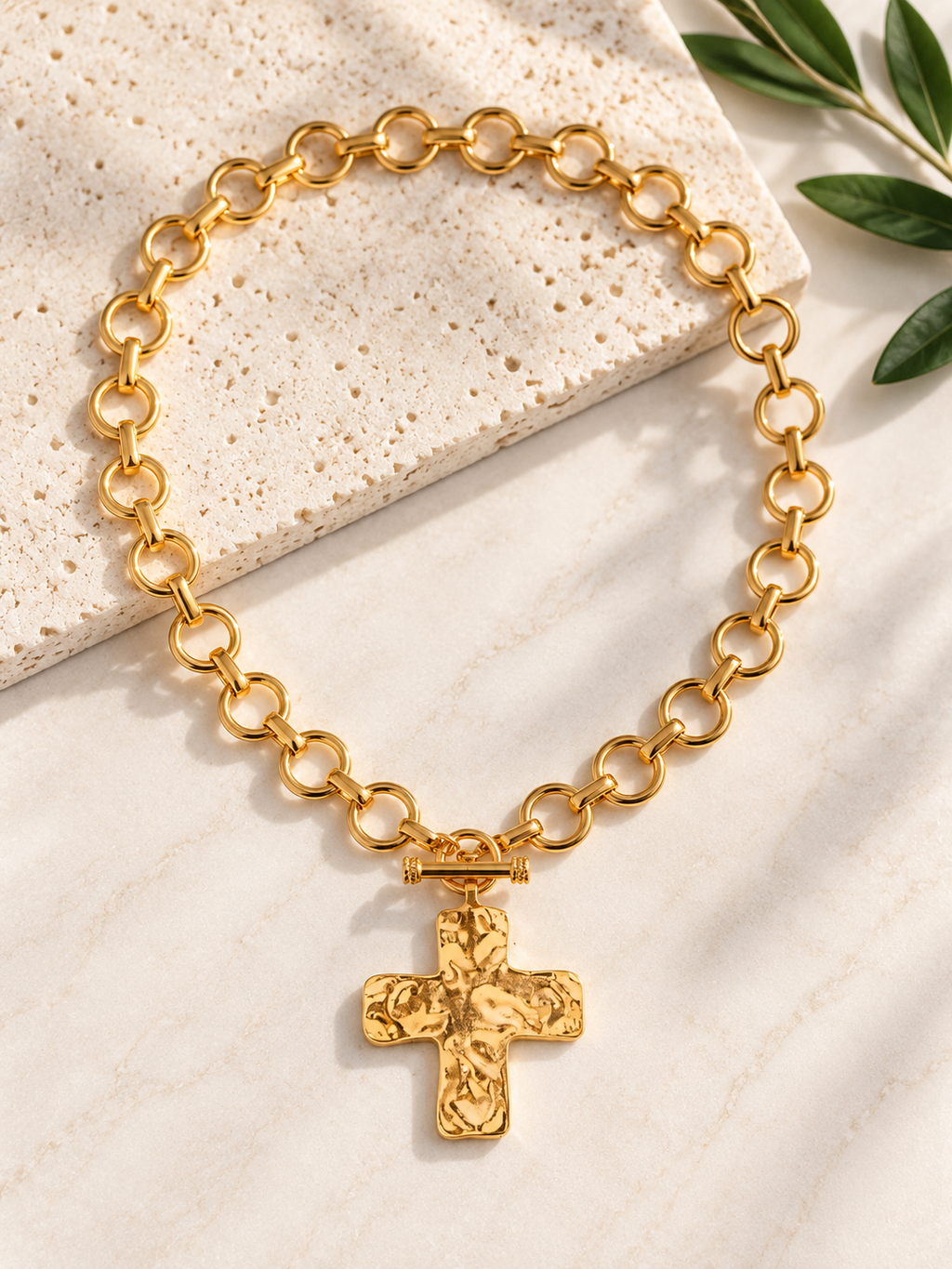 A gold chain necklace with a textured cross pendant, displayed on a light beige stone tile with green leaves in the corner.