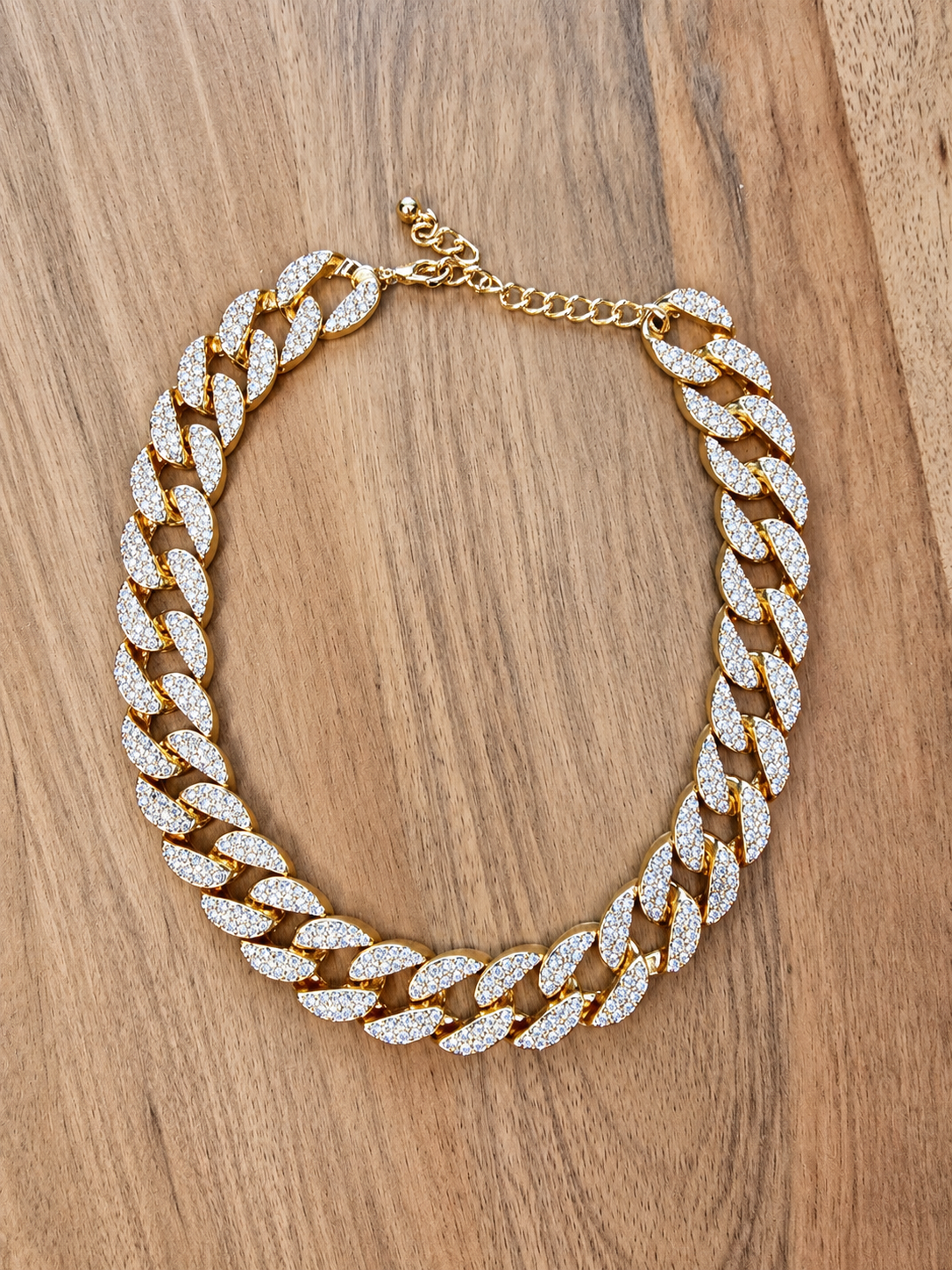 A chunky Cuban link chain necklace paved with clear crystals, displayed on a warm brown wooden surface.