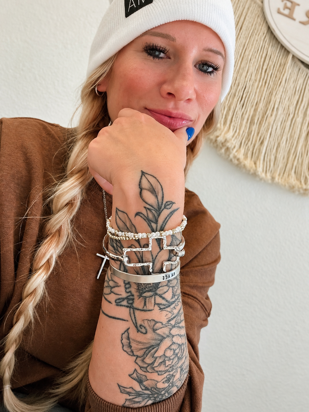 A close-up lifestyle shot of a woman with blonde hair in a braid, wearing a white beanie, brown top, and multiple silver bracelets on her wrist, resting her chin on her hand.