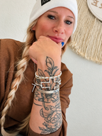 A close-up lifestyle shot of a woman with blonde hair in a braid, wearing a white beanie, brown top, and multiple silver bracelets on her wrist, resting her chin on her hand.