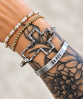 A close-up of a tattooed wrist wearing multiple bracelets, including a silver cross-shaped cuff with a lobster clasp, a silver bangle engraved with the word 'GRACE', and two beaded bracelets.