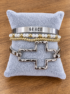 A stack of four bracelets displayed on a light blue plush jewelry pillow over a wooden surface. The bracelets include a wide silver cuff stamped with the word 'GRACE', a pearl and gold beaded bracelet, a gold and pearl beaded bracelet, and a textured silver cross-link bracelet.