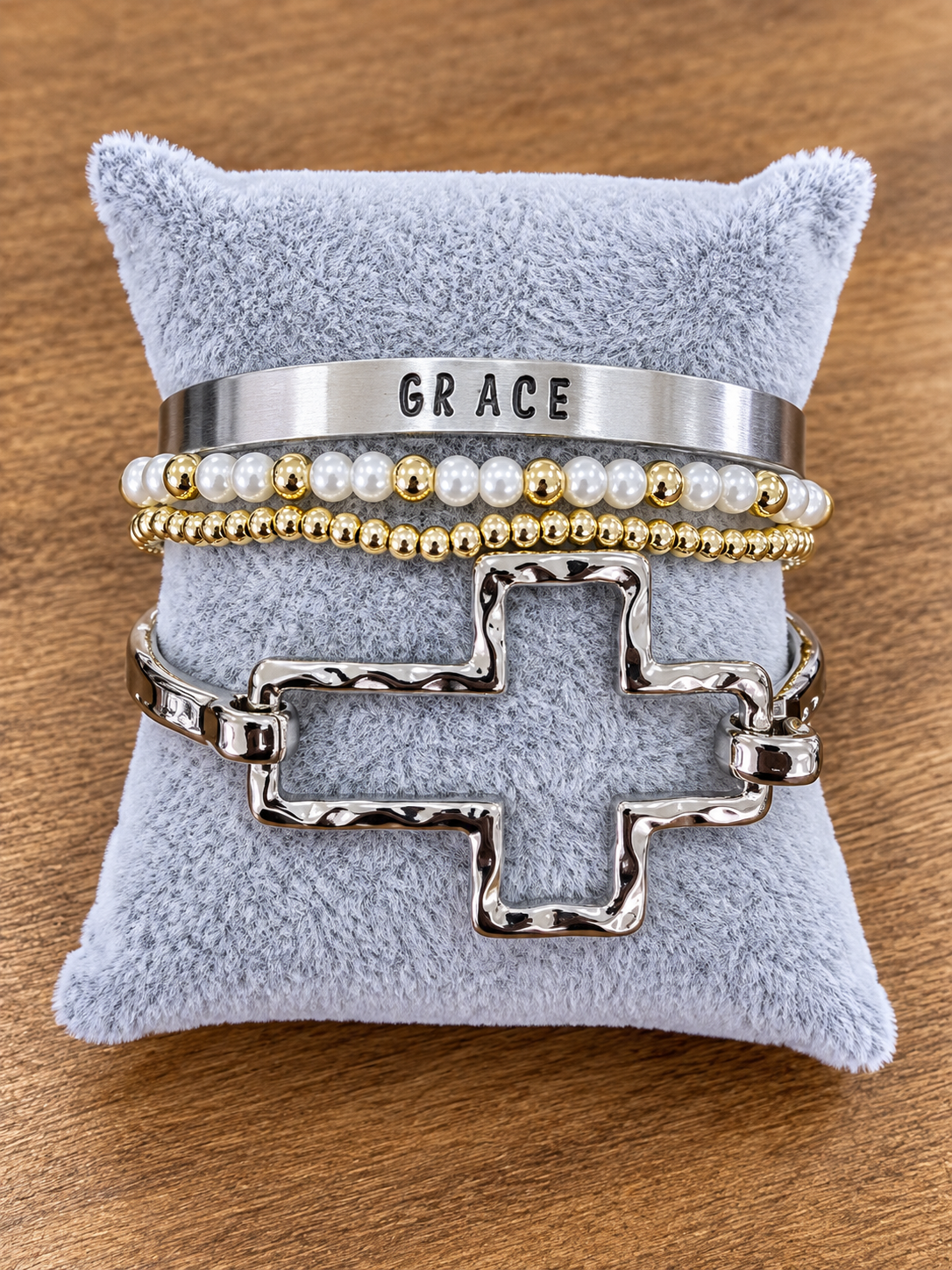 A stack of four bracelets displayed on a light blue plush jewelry pillow over a wooden surface. The bracelets include a wide silver cuff stamped with the word 'GRACE', a pearl and gold beaded bracelet, a gold and pearl beaded bracelet, and a textured silver cross-link bracelet.