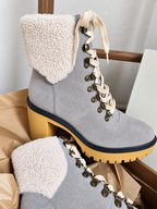 A pair of gray suede ankle boots with thick cream sherpa fleece cuffs, cream braided laces, and chunky yellow block heels, displayed on brown paper packaging.