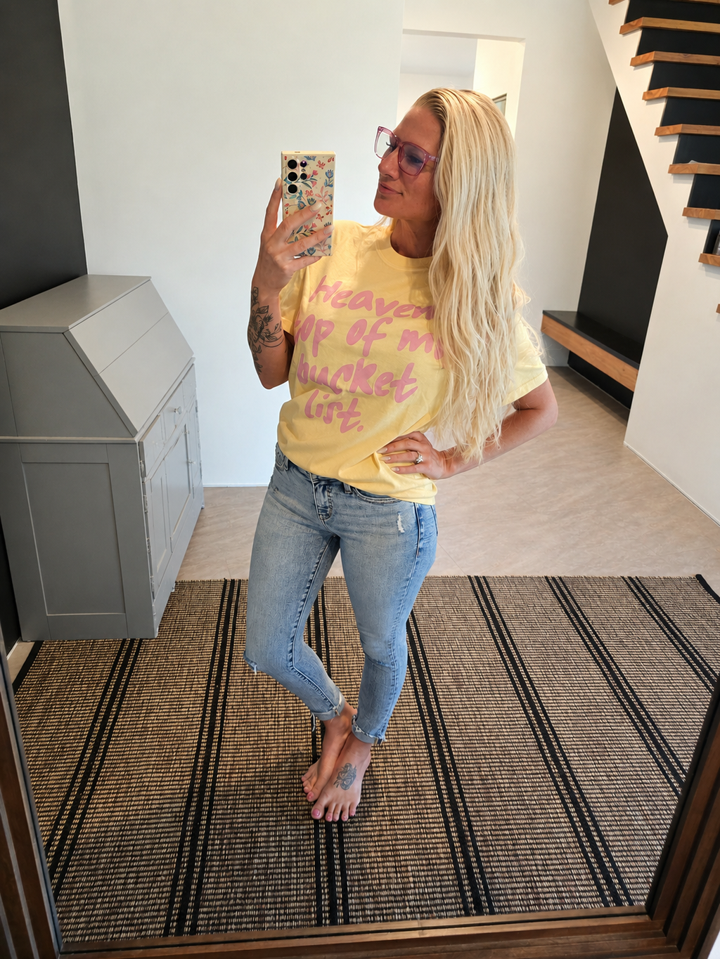 A woman takes a mirror selfie indoors wearing a yellow graphic T-shirt with pink text that reads 'Heaven is my bucket list' and light-wash blue jeans.