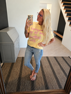 A woman takes a mirror selfie indoors wearing a yellow graphic T-shirt with pink text that reads 'Heaven is my bucket list' and light-wash blue jeans.