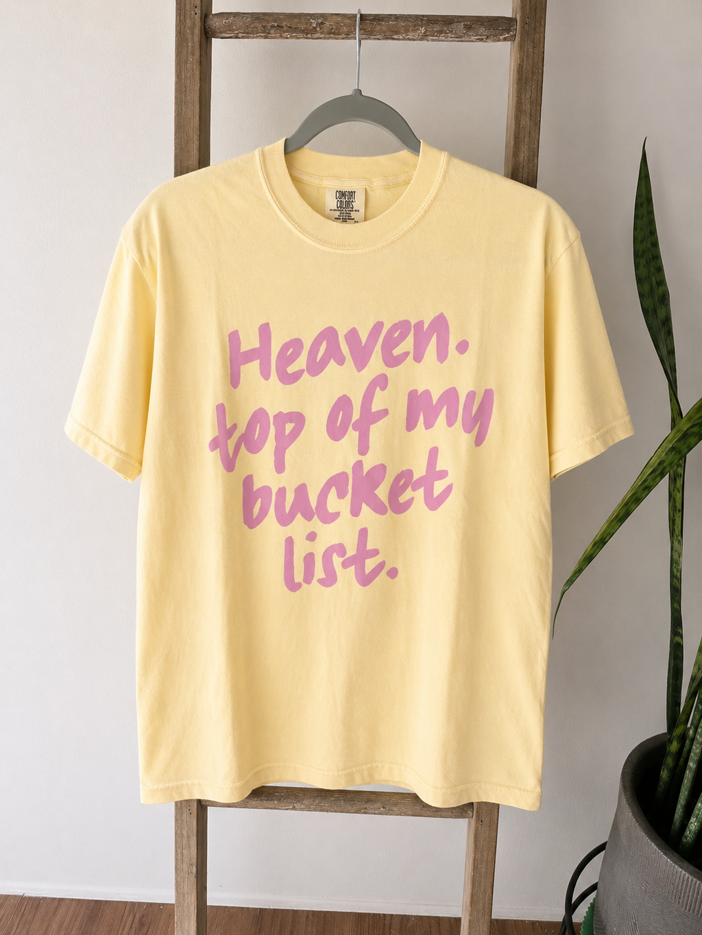 A pale yellow short-sleeve t-shirt hanging on a wooden ladder, featuring the phrase 'Heaven. top of my bucket list.