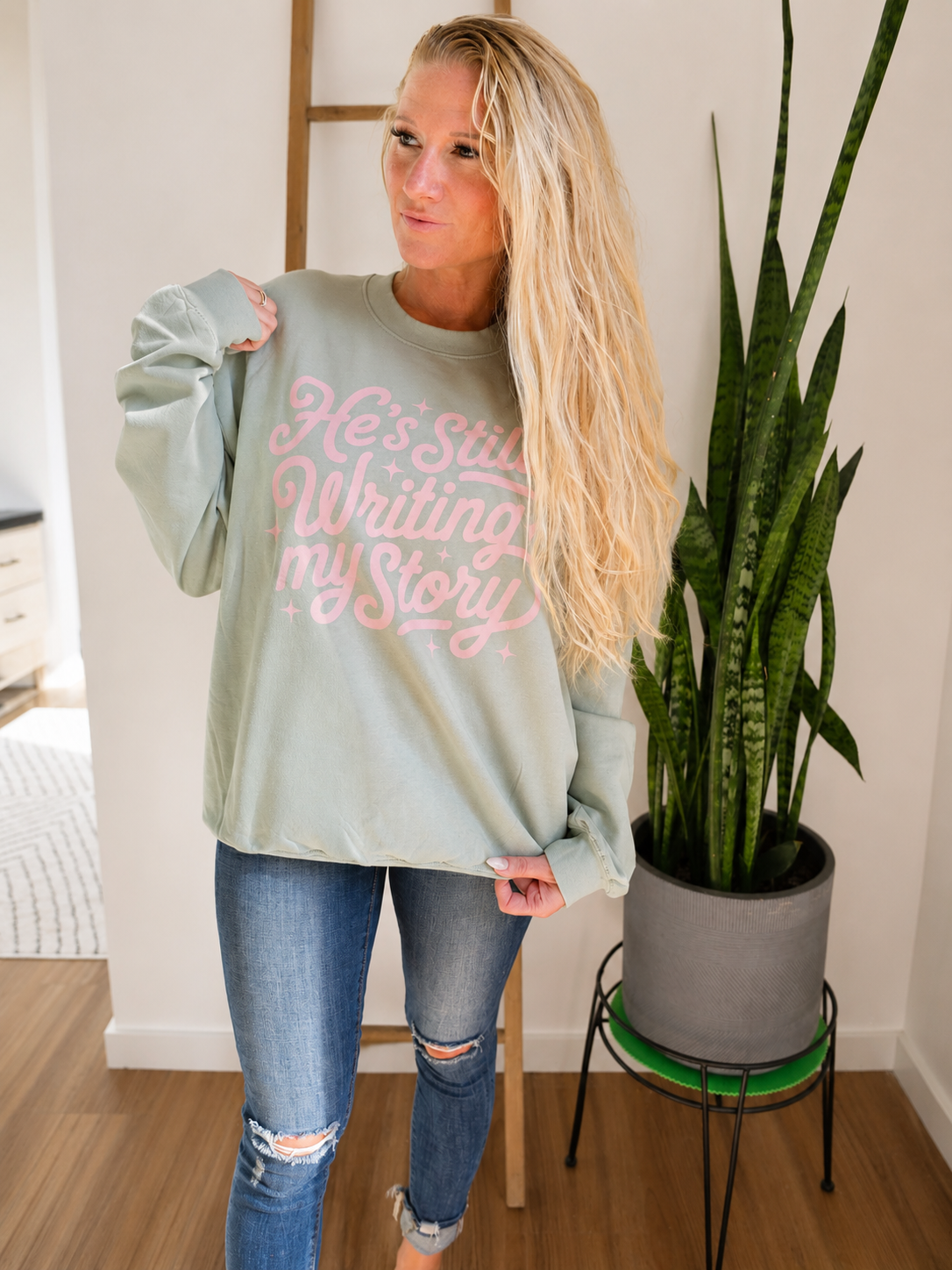 A woman wearing a sage green crewneck sweatshirt with pink cursive text that reads 'He's Still Writing My Story', paired with distressed blue jeans, standing in a room next to a potted snake plant.