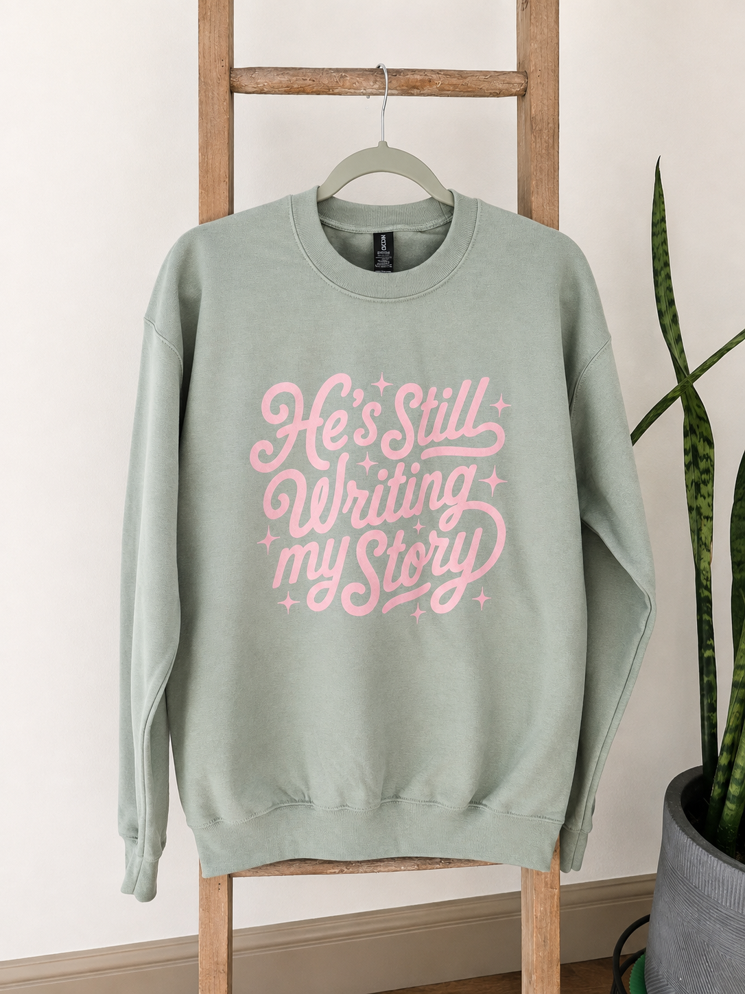 A sage green crewneck sweatshirt with a pink retro-style graphic reading 'He's Still Writing my Story' hanging on a wooden ladder against a white wall.