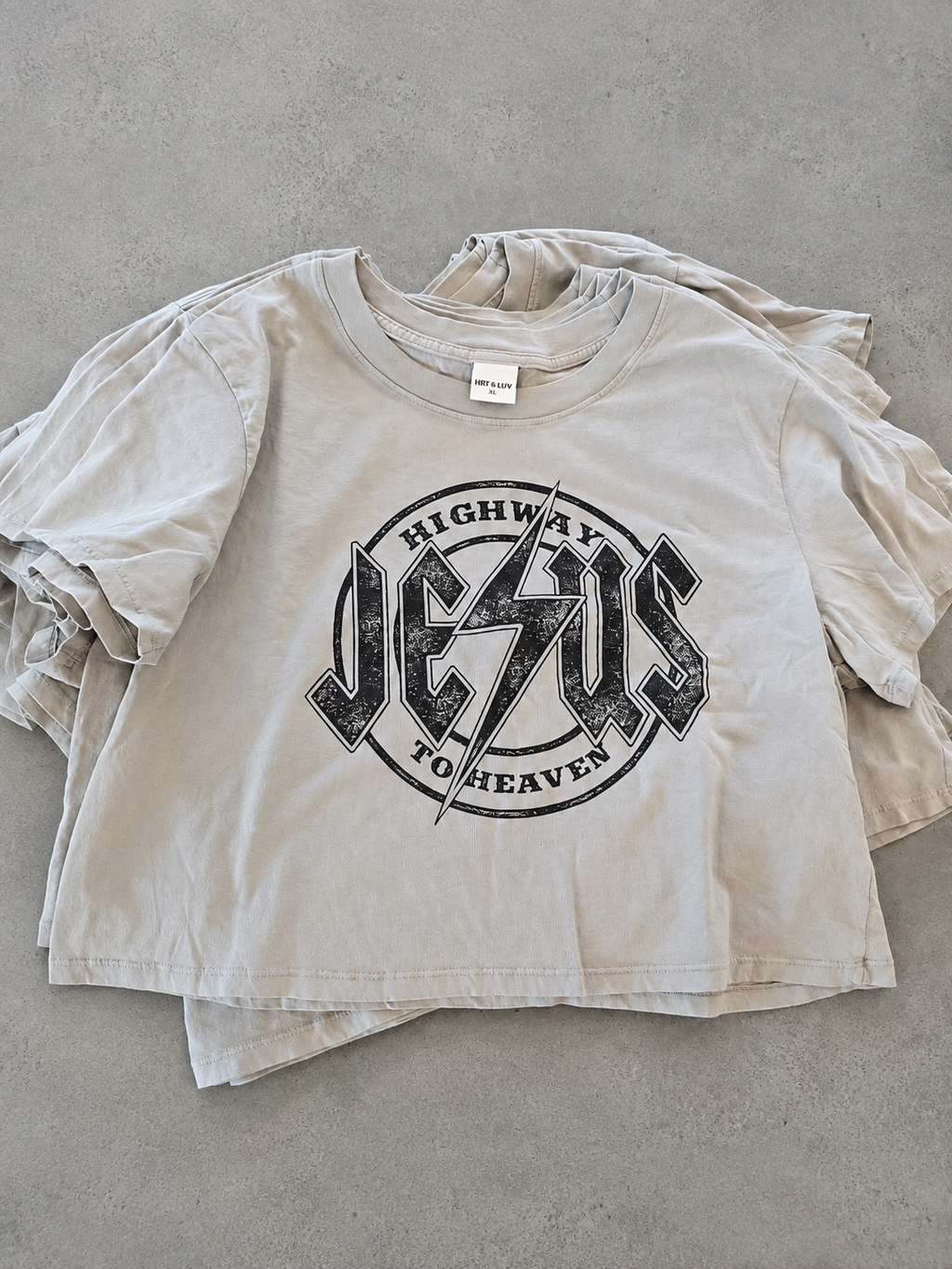 A light beige cropped t-shirt with a distressed black graphic reading 'HIGHWAY JESUS TO HEAVEN' on the front, laid flat on a gray concrete surface.