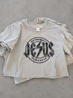 A light beige cropped t-shirt with a distressed black graphic reading 'HIGHWAY JESUS TO HEAVEN' on the front, laid flat on a gray concrete surface.