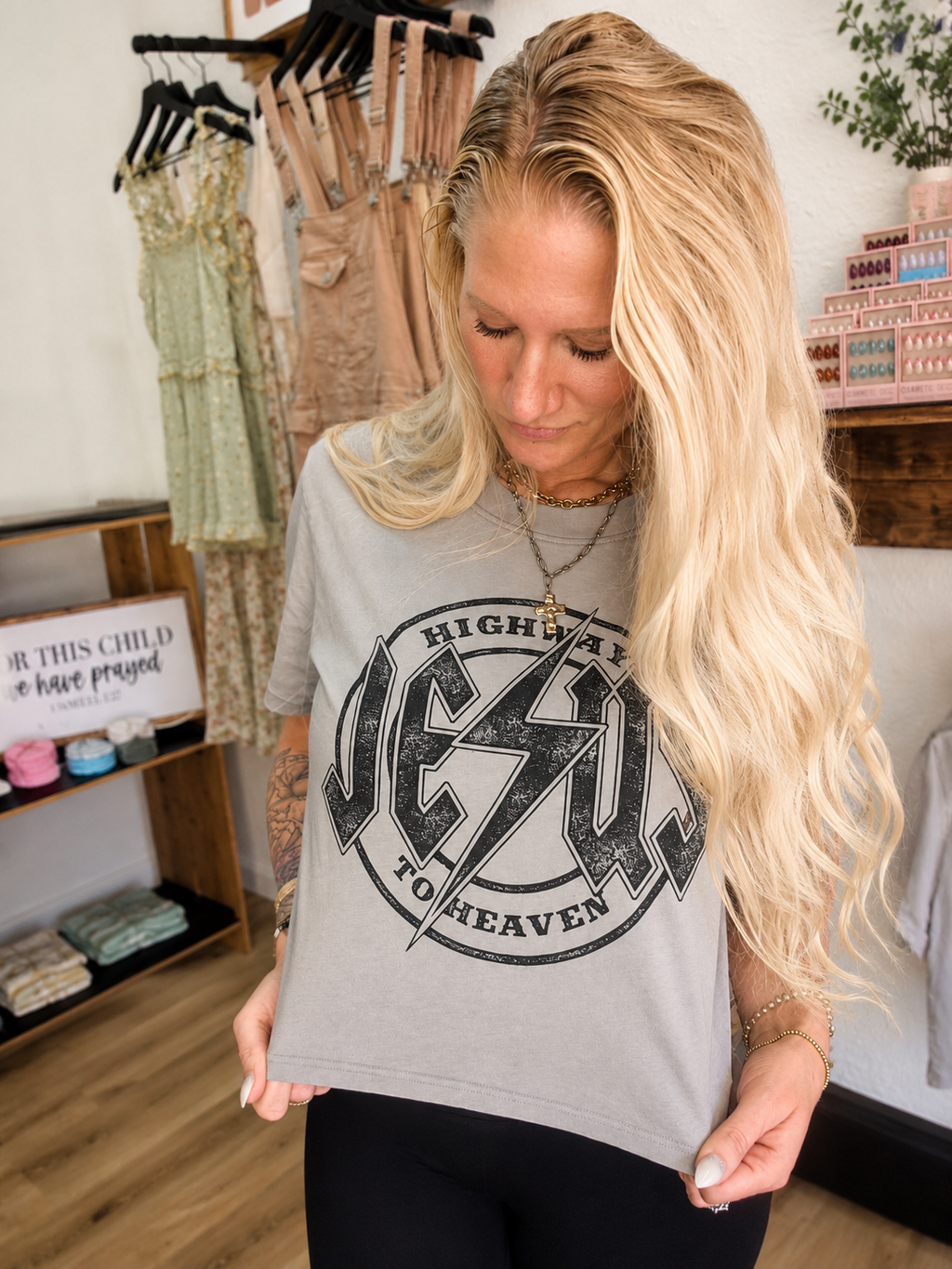 A woman with long blonde hair wearing a gray graphic t-shirt with a black distressed 'Jesus to Heaven' design, standing in a boutique setting.