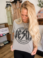 A woman with long blonde hair wearing a gray graphic t-shirt with a black distressed 'Jesus to Heaven' design, standing in a boutique setting.