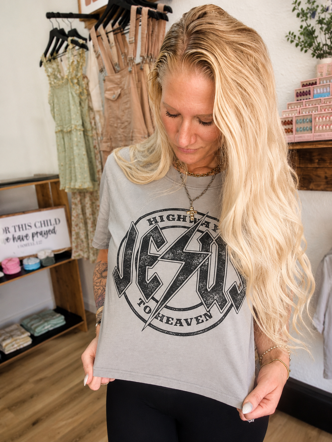 A woman with long blonde hair wearing a gray graphic t-shirt with a black distressed 'Jesus to Heaven' design, standing in a boutique setting.